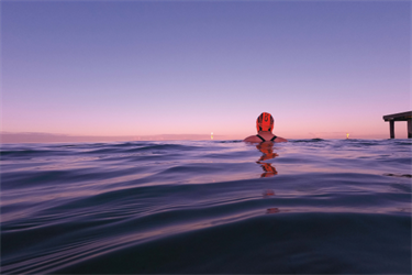 The back of a swimmer's head is visible above the water. The sky is pink and is reflecting on the surface of the water.