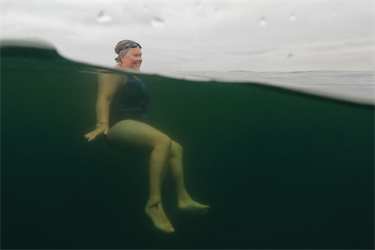 The camera is half-submerged in the water. A swimmer is seen, their head above the water and their body below. The water is dark green and the sky is cloudy.