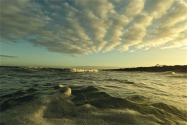 A view of the water and sky at the beach. The sky has rippling clouds, lit by the sun on the right-hand side of the image. The light reflects on the water.